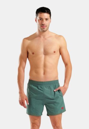 Uomo senza maglietta con capelli corti e scuri che indossa pantaloncini da bagno verdi con una vita elastica, in piedi con una mano in tasca davanti a uno sfondo uniforme.