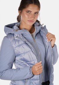 Giacca puffer azzurro chiaro con zip frontale, cappuccio e polsini elastici. Presenta un motivo trapuntato e dettagli in metallo dorato.