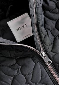 Sort quilted jakke indvendig med en sølvfarvet lynlås og et mærke med teksten "NEXT." Stoffet har en glat tekstur med et subtilt mønster.