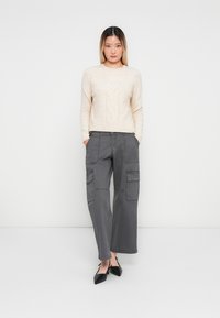 Donna in piedi con un maglione beige, pantaloni cargo grigi e scarpe nere a punta su uno sfondo bianco semplice. Mani nelle tasche.