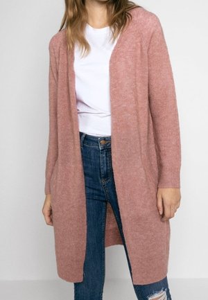 Kvinde iført en lang, åben lyserød cardigan over en hvid t-shirt og revnede blå jeans mod en simpel lys baggrund.