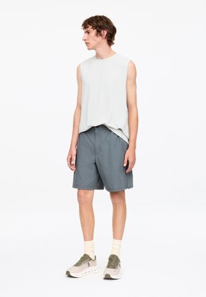 Jeune homme debout, portant un débardeur blanc, un short gris, des chaussettes claires et des chaussures de course blanches, regardant vers sa gauche.