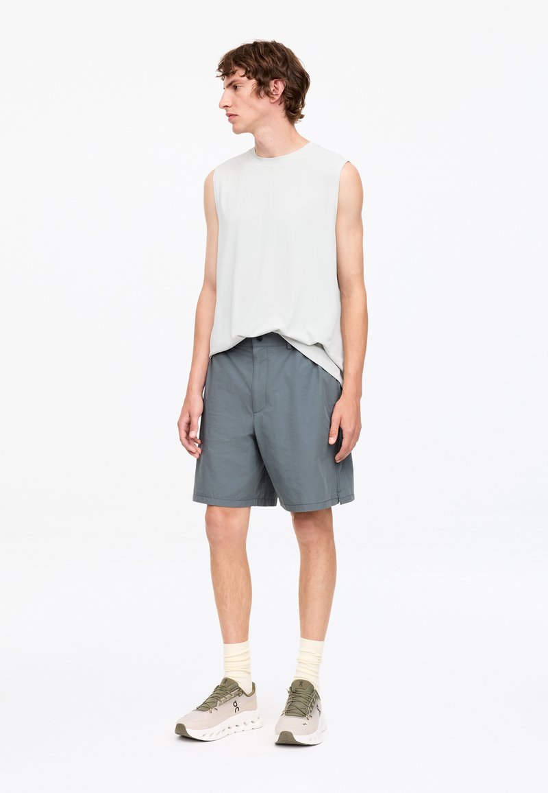 Jeune homme debout, portant un débardeur blanc, un short gris, des chaussettes claires et des chaussures de course blanches, regardant vers sa gauche.