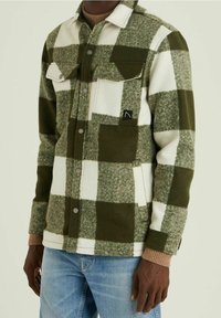 Homme portant une veste à carreaux verte et blanche boutonnée par-dessus un pull marron et un jean bleu clair, debout devant un fond uni.