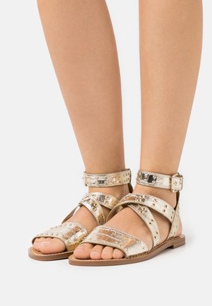 Metallische gouden sandalen met meerdere banden, een enkelgesp, studs en een platte zool. Open teen ontwerp toont de voet.