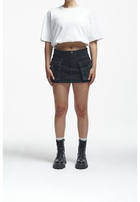 Jupe mini en denim noir avec poches avant, associée à un t-shirt blanc court et des bottes noires à lacets. Fond uni.