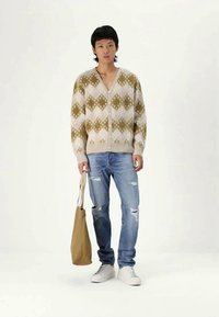 Cárdigan de color crema y verde oliva con un patrón y escote en V, combinado con jeans azules desgastados y zapatillas blancas, llevando un bolso de piel color marrón claro.