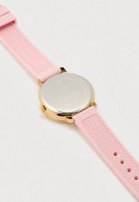 Guess polshorloge met goudkleurige kast, roestvrijstalen achterkant, en een gestructureerde roze siliconen band, plat liggend op een witte ondergrond.