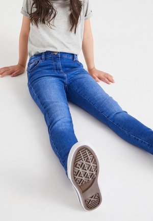 Enfant assis par terre portant un jean bleu, un t-shirt gris et des baskets blanches avec un motif de semelle visible.
