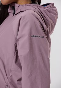 Ljust lila vattentät jacka med huva, med en slät yta och elastiska ärmslut. Logotypen 'rukka' tryckt i mörkblått på ärmen.
