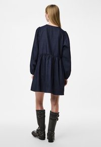 Vestido de ganga azul-escura com mangas longas e bufantes, cintura franzida e saia até ao joelho. Botas pretas até ao joelho com detalhes em fivela completam o visual.
