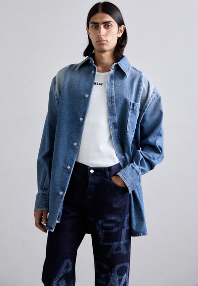 Camică din denim cu un gradient de culoare albastră care trece de la deschis la închis. Are o buzunar și nasturi. Asortată cu blugi negri care prezintă un model grafic.