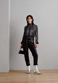 Blouse noire à motifs avec col haut et manches longues, associée à un pantalon en cuir noir et des bottines à talons argentés. Tenant un petit sac noir.
