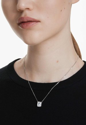 Collier en chaîne argentée avec un pendentif rectangulaire transparent, porté sur un pull noir, mettant en valeur son design simple et élégant.