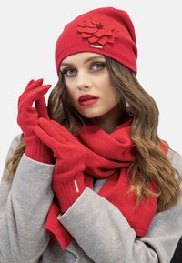 Czerwony dzianinowy beanie z filcowym detalem w kształcie kwiatu, współgrające czerwone rękawiczki i szalik. Modelka nosi szary płaszcz, który eksponuje teksturowane materiały i żywe kolory.