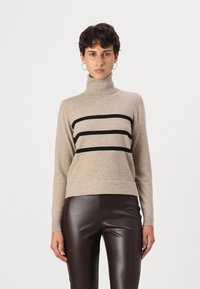 Beige garbó, amelyen három vízszintes fekete csík található, hosszú ujjaival és enyhén cropped nyakrésszel, barna bőr nadrággal párosítva.