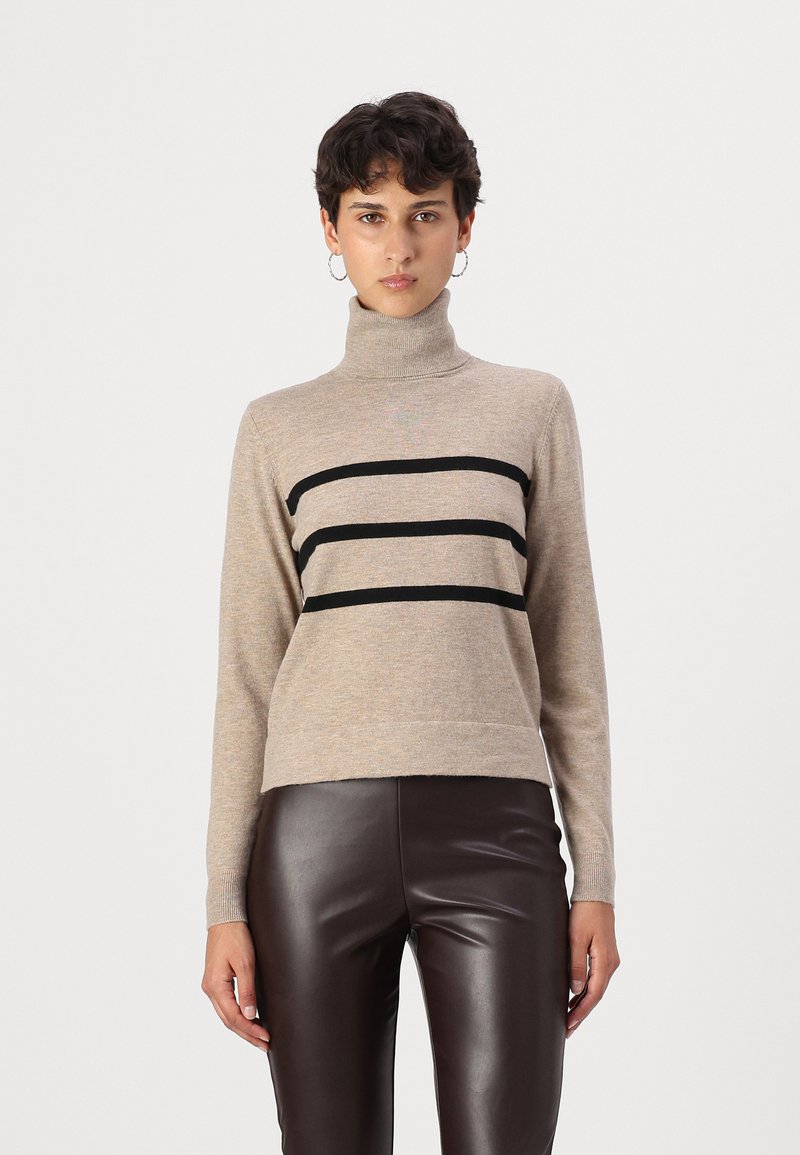 Beige garbó, amelyen három vízszintes fekete csík található, hosszú ujjaival és enyhén cropped nyakrésszel, barna bőr nadrággal párosítva.