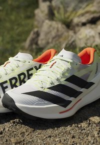 Ζευγάρι λευκά παπούτσια Adidas Terrex για ορεινό τρέξιμο με μαύρες ρίγες, κορδόνια νέον κίτρινου χρώματος και πορτοκαλί εσωτερική επένδυση τοποθετημένα σε πετρώδες έδαφος σε εξωτερικό χώρο.