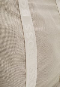 Borsa in tessuto beige strutturato con una fascia bianca in rilievo ben visibile, con il nome del marchio "BOSS" ripetuto lungo il lato.