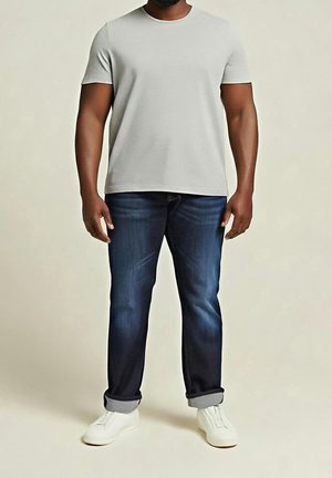 Homme portant un t-shirt gris clair uni à manches courtes, un jean bleu foncé retroussé et des baskets blanches, debout devant un fond neutre.