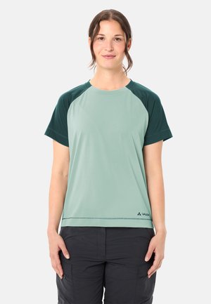 Donna in piedi, rivolta verso la fotocamera, che indossa una maglietta a maniche corte verde chiaro e verde scuro e pantaloni cargo neri, su sfondo uniforme.