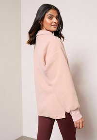 Roosa fliispullover, millel on krae, pikad varrukad ja pehme tekstuur. Seljas on bordokarva retuusid. Ühtlane taust.