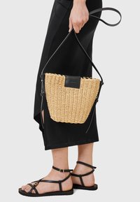 Borsa a secchiello in paglia intrecciata con un accento in pelle nera, dotata di una lunga tracolla regolabile. Superficie testurizzata e forma circolare.