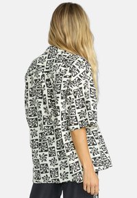 Camicia bianca con grande motivo floreale nero, maniche corte, vestibilità ampia e dettagli di design quadrato. La vista dal retro mostra una silhouette rilassata.