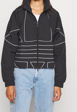 Mujer con chaqueta negra con cremallera y líneas geométricas blancas y vaqueros de talle alto color azul claro sobre un fondo liso.