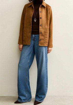 Vrouw met een bruine jas met knopen, een donkerbruine coltrui, brede blauwe jeans en puntige donkere schoenen tegen een effen achtergrond.