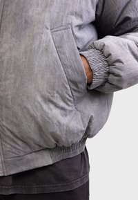 Gray puffer jacket with a matte finish, elastic cuffs, side pocket, and ribbed hem, featuring a soft, textured appearance.