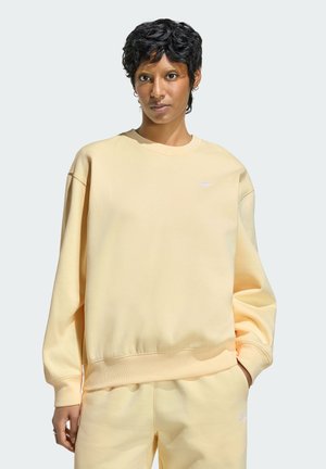 Donna con capelli neri corti e ricci che indossa una felpa oversize giallo chiaro e pantaloni abbinati, in piedi con una mano in tasca.
