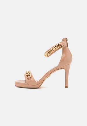 Beige høje hæle sandal med åben tå, ankrem og guldkæde dekoration på begge remme, sat mod en ensfarvet hvid baggrund.