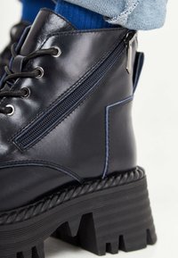 Stivaletto alla caviglia in pelle nera con plateau testurizzato, dotato di occhielli argentati, lacci, una cerniera laterale e cuciture a contrasto blu.