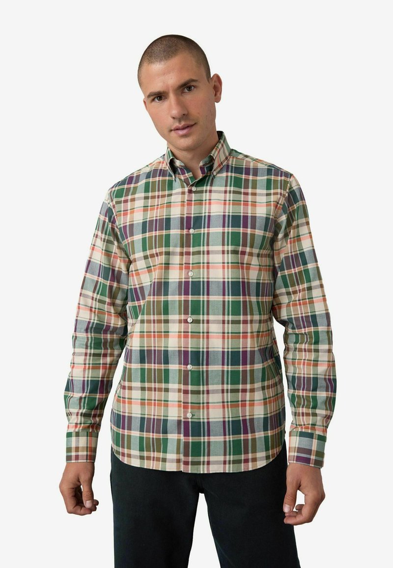 Man met kort haar die een multi-kleurig ruitjesoverhemd met knopen draagt. Hij staat met een ontspannen gezicht tegen een effen witte achtergrond.
