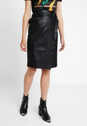 Kobieta w czarnej skórzanej ołówkowej spódnicy z kopertowym zapięciem, czarnych botkach za kostkę z metalowymi noskami oraz czarnej koszulce z grafiką.