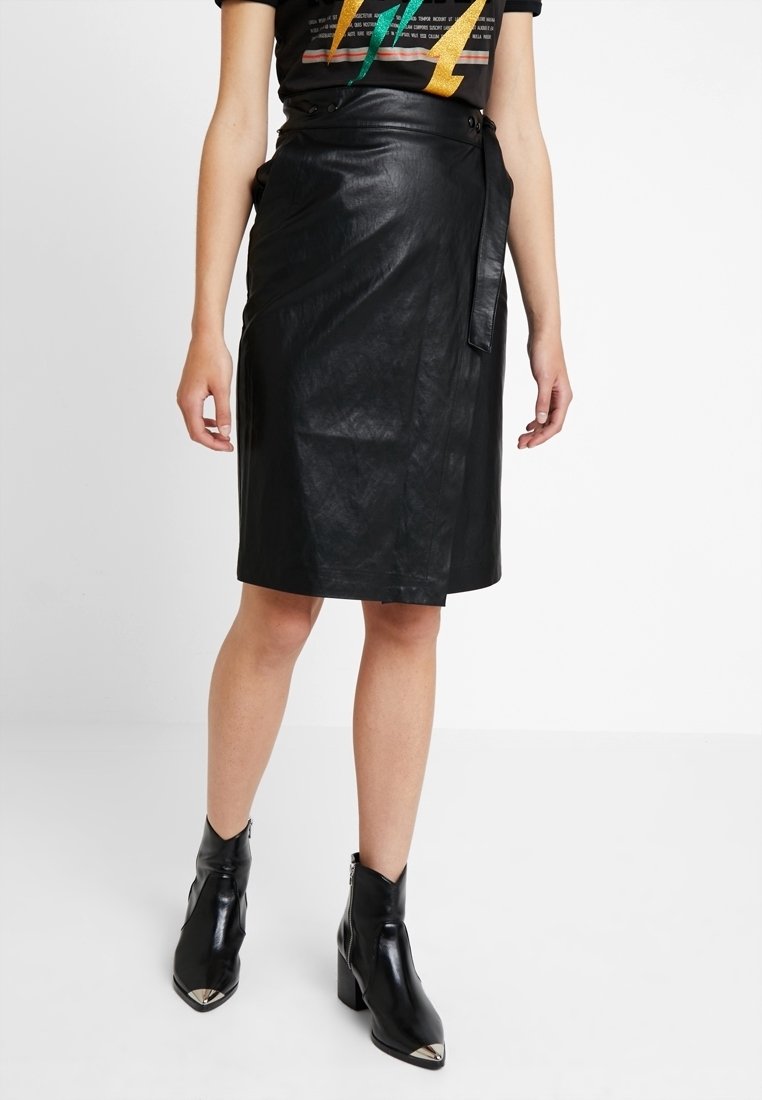 Femme portant une jupe portefeuille en cuir noir, des bottines noires à bouts métalliques et un t-shirt noir à imprimé graphique.