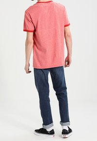 Camiseta roja de manga corta con cuello y puños de canalé, combinada con jeans de mezclilla oscura, recogidos a los tobillos, y zapatillas negras.