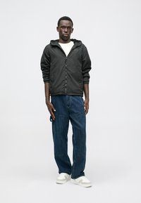 Svart huva jacka med dragkedja, bärs över en vit skjorta. Blå denimjeans och vita sneakers fullbordar outfiten. En enkel bakgrund.