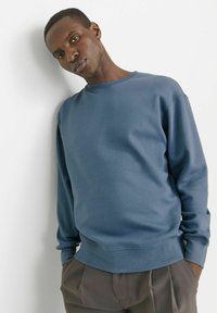 Sudadera azul de cuello redondo hecha de una tela suave, con mangas largas, puños acanalados y un corte relajado, combinada con pantalones grises.