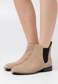 Botines de ante beige con textura suave, paneles laterales elásticos negros y una suela plana con un ribete decorativo de cuentas doradas.