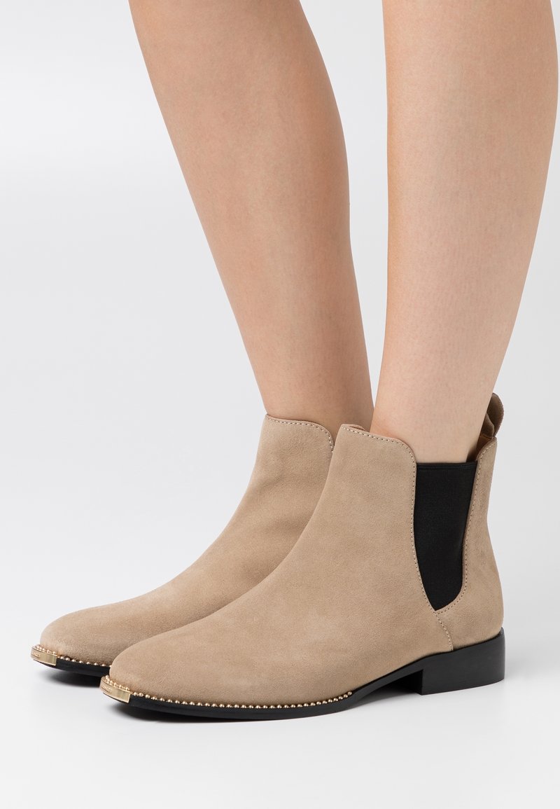 Botines de ante beige con textura suave, paneles laterales elásticos negros y una suela plana con un ribete decorativo de cuentas doradas.