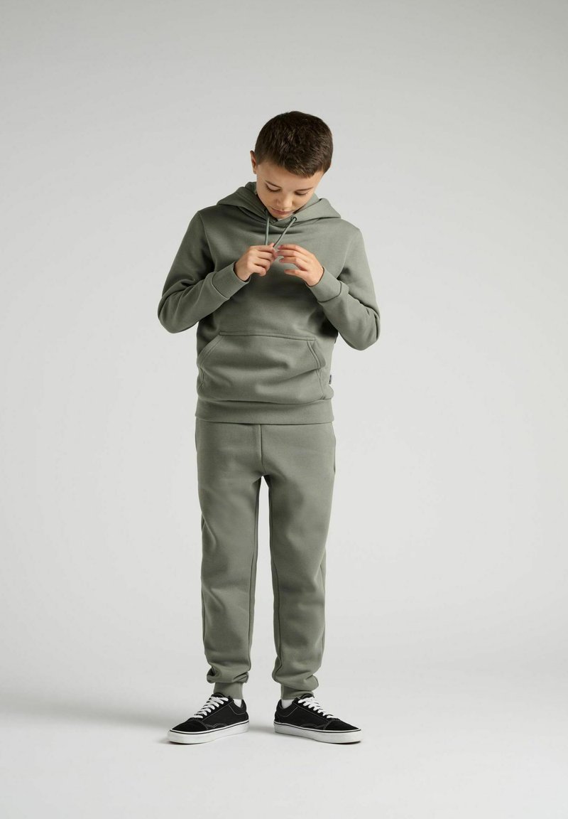 Olivengrøn hætte sweatshirt med snore, frontlomme, matchende joggers og sorte sneakers med hvid sål. Blødt materiale med glat tekstur.