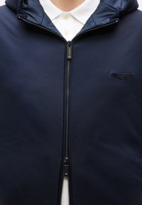 Σκούρο μπλε μπουφάν με φερμουάρ πάνω από ένα λευκό πουκάμισο με γιακά, με ανασηκωμένο μαύρο έμβλημα με φτερά στο αριστερό στήθος.