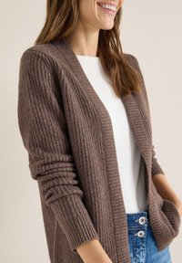 Hnědý pletený cardigan s žebrovanou texturou, otevřený vpředu, s dlouhými rukávy, kombinovaný s bílým tričkem a modrými džínami z denimu.