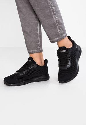 Person iført sorte Skechers sportssko og grå bukser, der står på en lysegrå gulv med den ene fod let hævet.