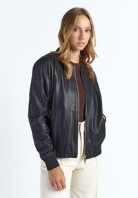 Marineblå bomberjakke i læder med ribstrikkede manchetter og kant, forsynet med en gylden lynlås og sidelommer, båret over en brun top og cremefarvede bukser.