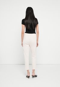 Schwarzes, eng anliegendes T-Shirt, hellrosa maßgeschneiderte Hosen mit elastischem Bund und schwarze, offene Absatzmules. Hintergrund ist einfarbig weiß.