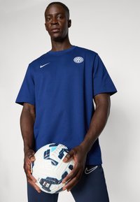 Sötétkék pamut póló fehér logóval és Nike pipa emblémával, sötét tengerészkék nadrággal párosítva. A modell egy fehér focilabdát tart, kék díszítésekkel.