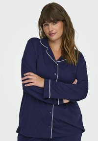 Mörkblå pyjamasset i mjukt material med vita detaljer, som har en knapp-up skjortdesign och långa ärmar. Modellen poserar med armarna i kors.